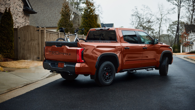 A burnt orange Toyota Tacoma TRD Pro parked on a residential street, featuring a bike rack mounted in the truck bed, surrounded by houses and trees on an overcast day.