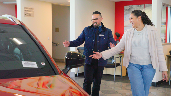 A car salesperson in a navy jacket and glasses enthusiastically shows a red vehicle to a smiling woman in a cream sweater and jeans inside a dealership.