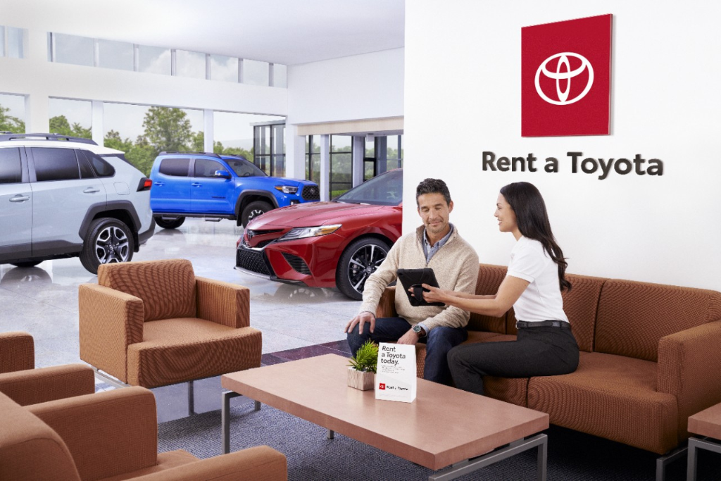 A Toyota Vallejo rental associate shows a customer a tablet in a dealership lounge with a “Rent a Toyota” sign on the wall and three Toyota vehicles—an SUV, truck, and sedan—visible through the showroom windows.