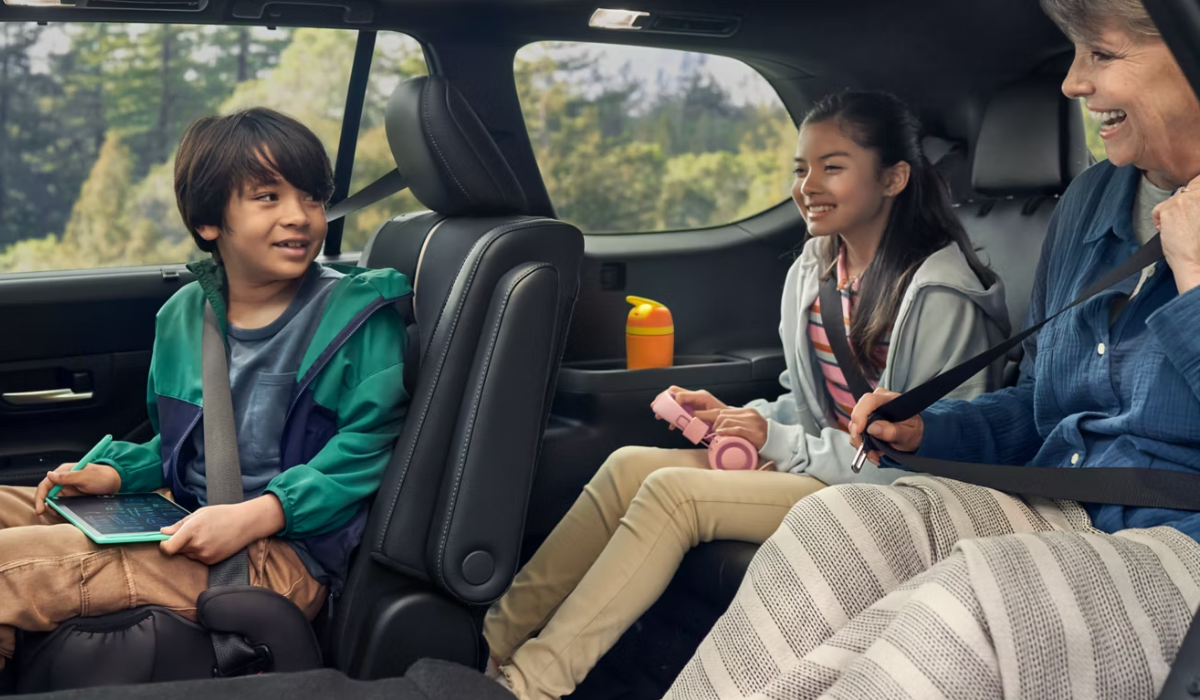 Two children and an older woman smiling and talking while riding in the back seat of a car, all wearing seat belts.