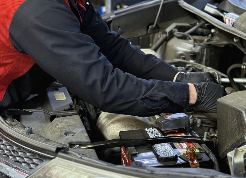 Hands in black gloves work on a car battery in an open engine compartment.
