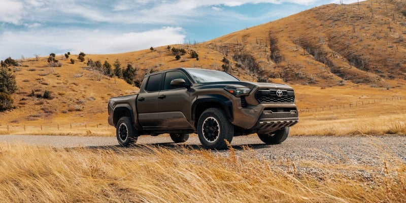 A dark green Toyota Tacoma truck parked on a gravel road in a vast, dry, hilly landscape under a blue sky.