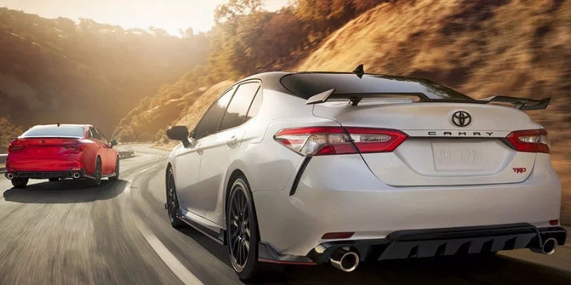 A white Toyota Camry TRD with a black spoiler leads a red Camry TRD on a winding road at sunset.